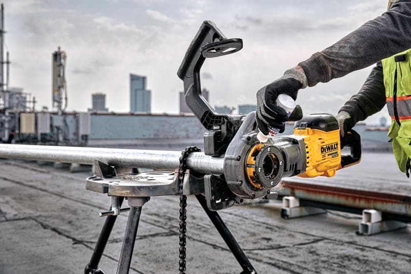 Eine Person mit Handschuhen und Sicherheitsweste bedient ein Elektrowerkzeug der Marke DEWALT, um ein Metallrohr zu schneiden oder zu gewindeschneiden, das in einem Dreibeinständer auf einer Industrie-Baustelle im Freien mit Stadtgebäuden im Hintergrund gehalten wird.