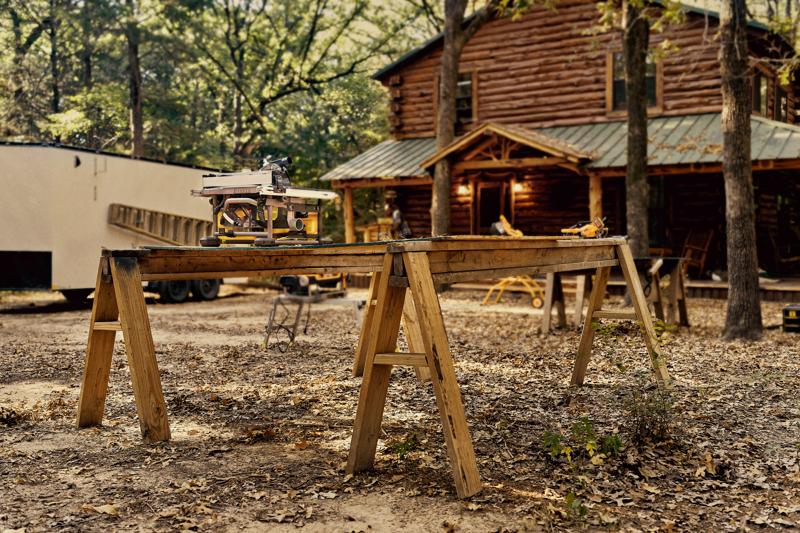 Eine DEWALT Tischkreissäge steht auf einer Holzwerkbank aus zwei Sägeböcken im Freien, im Hintergrund eine Blockhütte und Bäume.